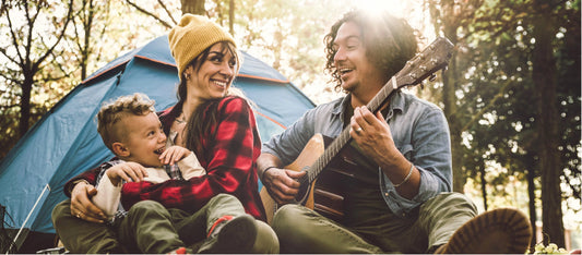 A family outdoor camping, one playing guitar as two others looks in his direction.